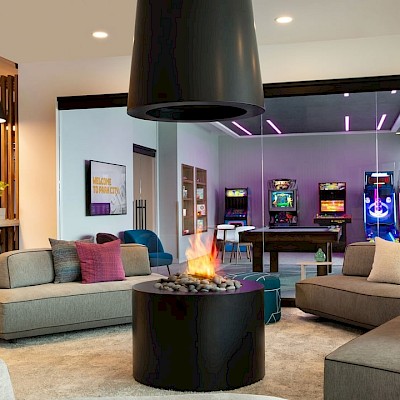 A modern lounge with cozy seating, a central fireplace, and an arcade area with games in the background, creating a vibrant atmosphere.