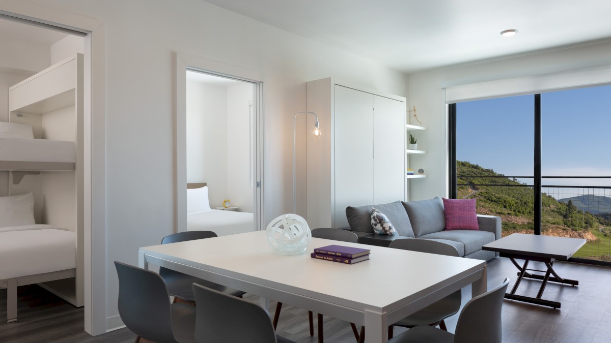 A modern living space with a white dining table, grey chairs, sofa, and glass doors opening to a scenic view; bunk beds in a nook.