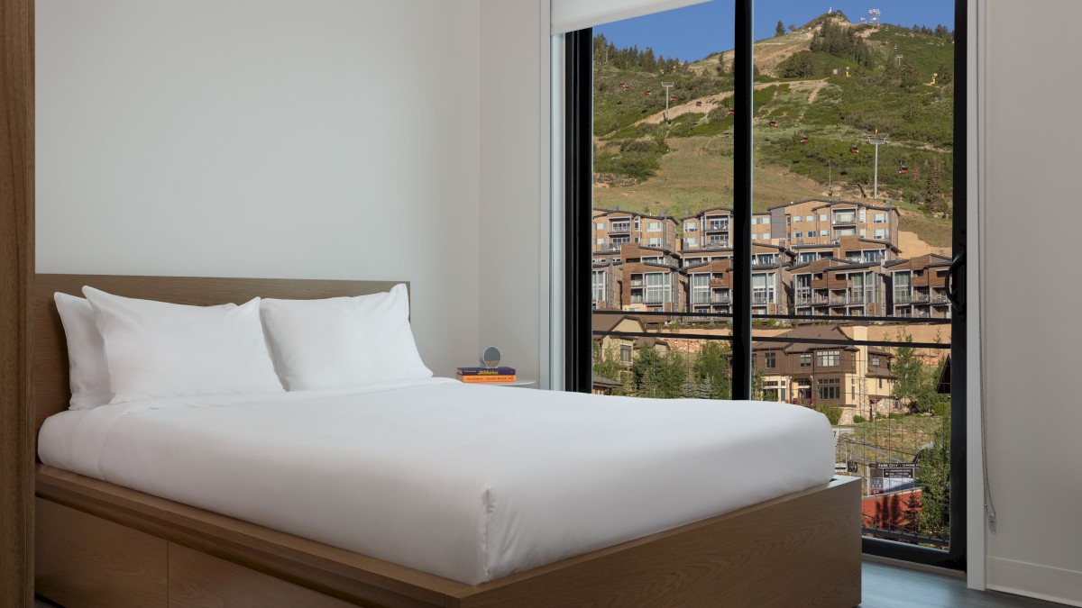 A cozy bedroom with a modern wooden bed and white bedding, overlooking a scenic hillside and buildings through a large window.