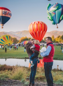 A family with a child and baby enjoy a hot air balloon festival, surrounded by colorful balloons and a scenic backdrop.