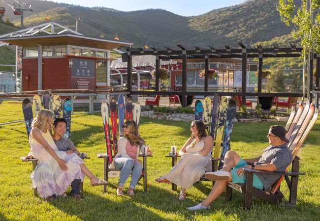 People sit outdoors on ski-themed chairs, chatting and laughing in front of a scenic mountain backdrop.