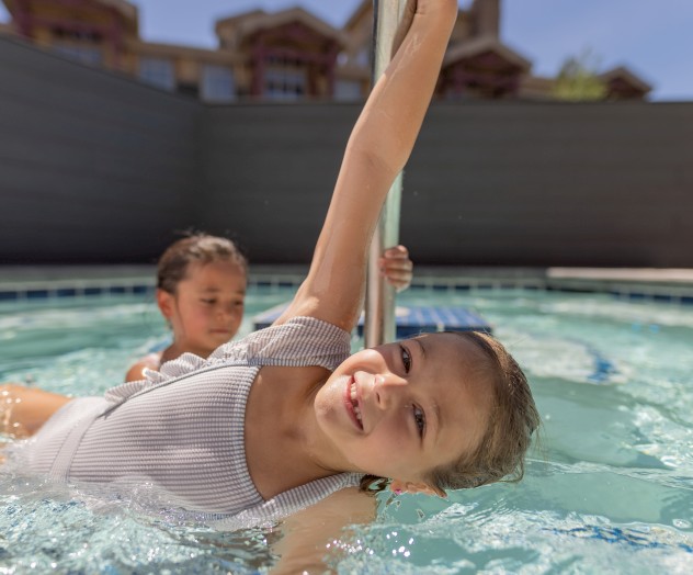 Two children are playing and smiling in a swimming pool, with one holding onto a metal pole in the water.