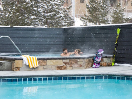 Two people relaxing in a hot tub surrounded by snow, with skis and a snowboard nearby; an outdoor pool is in the foreground.