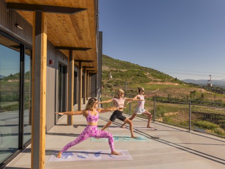 Three people are practicing yoga on a balcony with mats, overlooking a scenic landscape and ski lift in the background.
