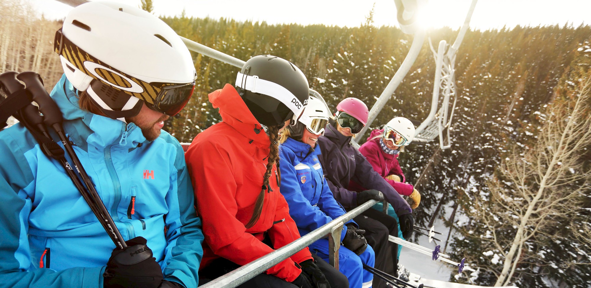 Five people wearing ski gear are sitting on a chairlift, surrounded by snowy trees and mountains.