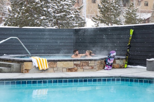 A couple relaxes in a hot tub surrounded by snow, with skis and a snowboard nearby, adjacent to a pool.