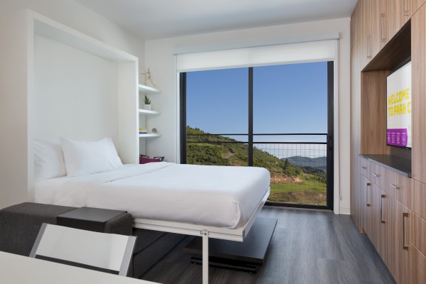 A modern hotel room featuring a bed, shelves, TV, and a large window with a scenic mountain view.