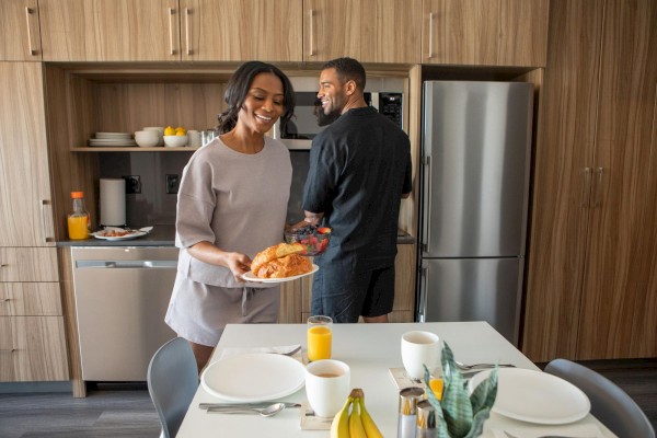 A couple is in a kitchen; one carries croissants, the other prepares fruit. The table is set with drinks and a plant centerpiece.