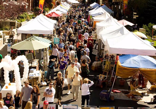 A bustling outdoor market with white tents, vendors, and visitors browsing the stalls in a lively atmosphere.
