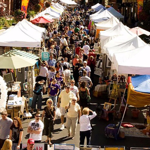 A bustling outdoor market with white tents, vendors, and visitors browsing the stalls in a lively atmosphere.
