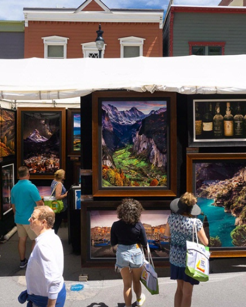 Outdoor art exhibit with paintings, people browsing, and sunny weather create a lively street scene.