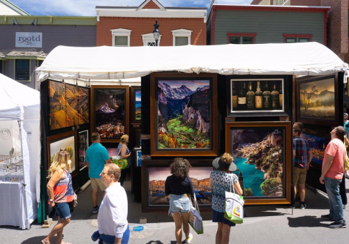 Outdoor art exhibit with paintings, people browsing, and sunny weather create a lively street scene.