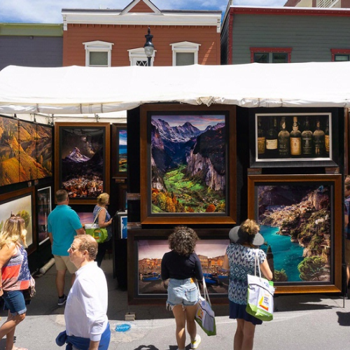 Outdoor art exhibit with paintings, people browsing, and sunny weather create a lively street scene.