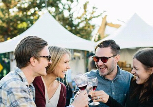 A group of four people are outdoors, happily clinking wine glasses together in front of white tents.