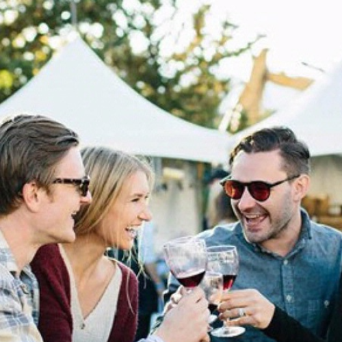 A group of four people are outdoors, happily clinking wine glasses together in front of white tents.
