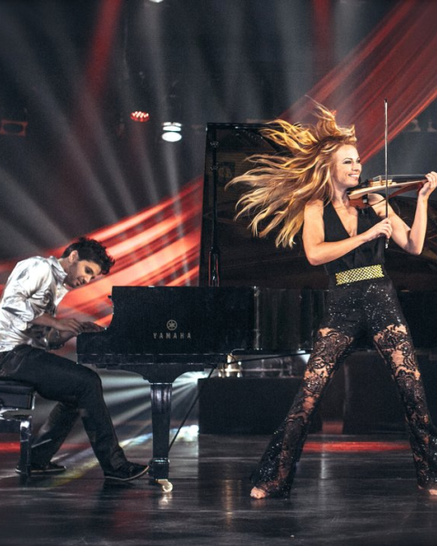 A dynamic performance with a pianist and a violinist on stage, surrounded by dramatic lighting and red drapes, create a vibrant atmosphere.