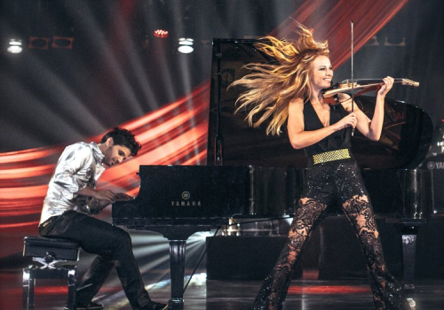 A dynamic performance with a pianist and a violinist on stage, surrounded by dramatic lighting and red drapes, create a vibrant atmosphere.