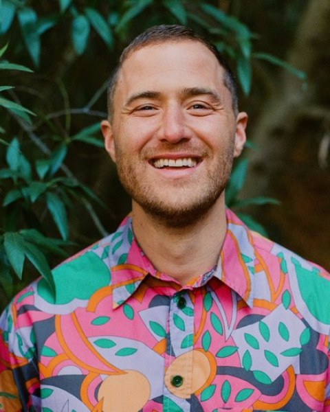 A person in a colorful, patterned shirt is smiling while standing near green foliage.