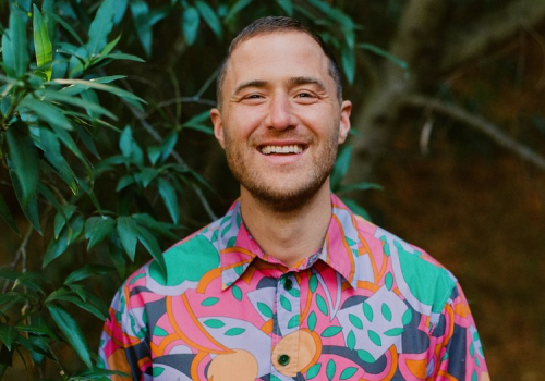 A person in a colorful, patterned shirt is smiling while standing near green foliage.