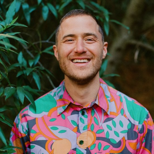 A person in a colorful, patterned shirt is smiling while standing near green foliage.