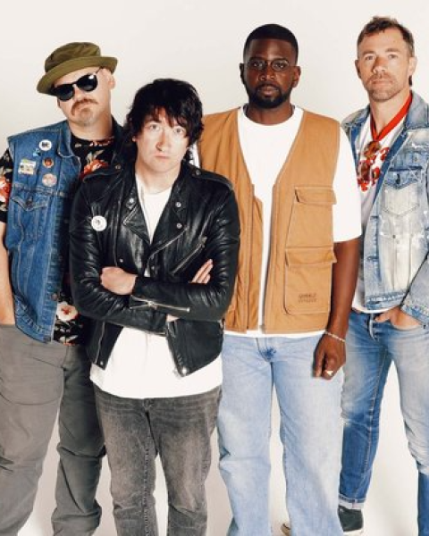 Four individuals in varied casual clothing, including denim and leather, stand against a plain backdrop.