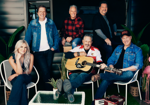 A group of six people sitting, two with guitars, smiling in a casual, outdoor setting with plants around them.