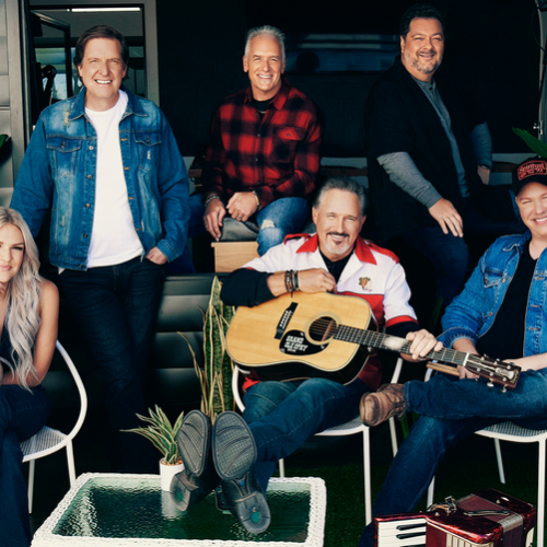 A group of six people sitting, two with guitars, smiling in a casual, outdoor setting with plants around them.