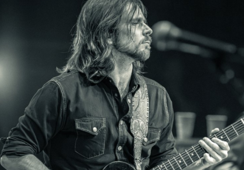 A person with long hair is playing an electric guitar on stage, with a microphone nearby, in a black and white setting.