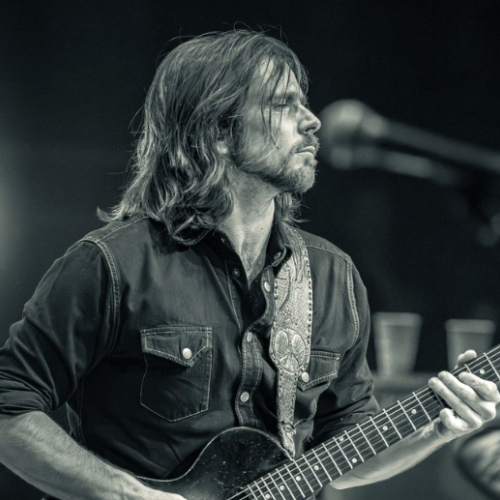 A person with long hair is playing an electric guitar on stage, with a microphone nearby, in a black and white setting.