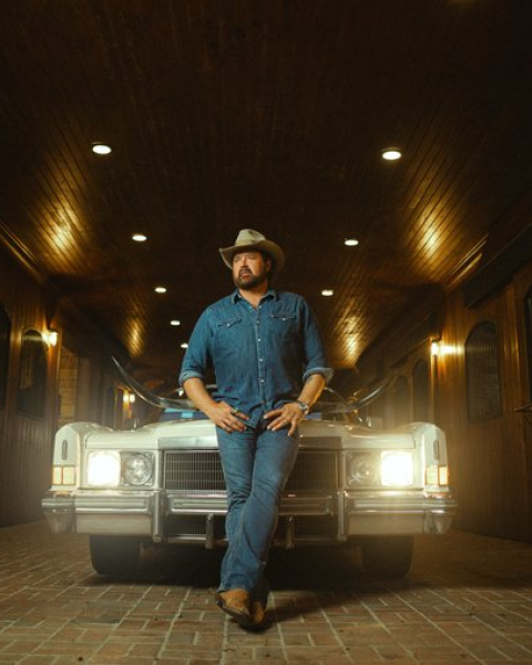 A person in cowboy attire leans against the front of a vintage car in a dimly lit, wood-paneled garage.