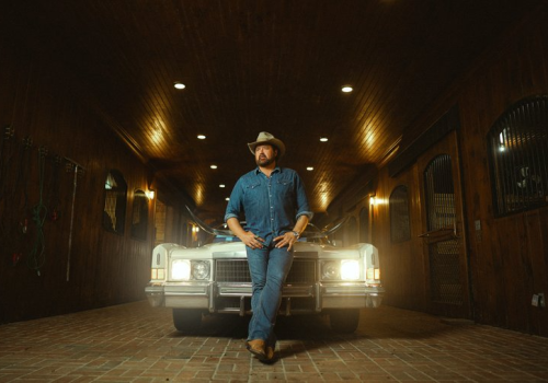 A person in cowboy attire leans against the front of a vintage car in a dimly lit, wood-paneled garage.