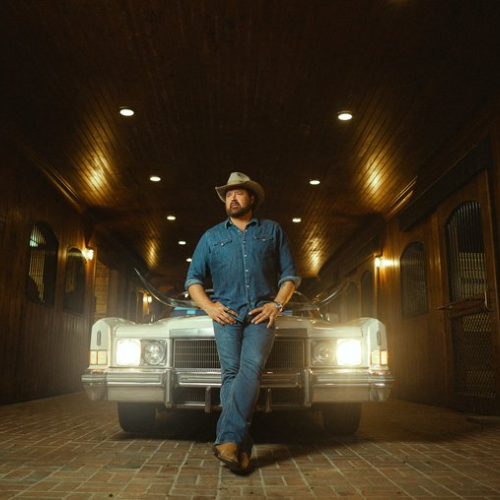 A person in cowboy attire leans against the front of a vintage car in a dimly lit, wood-paneled garage.