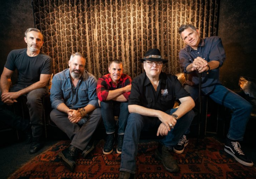 Five men are sitting together in a relaxed, casual setting with a patterned backdrop.