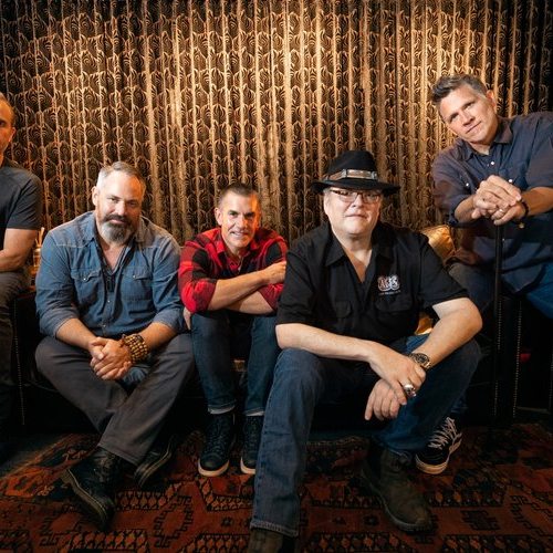 Five men are sitting together in a relaxed, casual setting with a patterned backdrop.