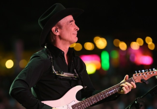 A person in a black outfit and hat is playing an electric guitar with a harmonica holder, against a colorful, blurred lights background.