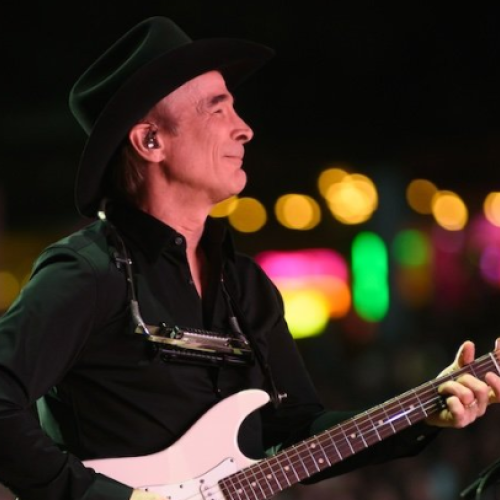 A person in a black outfit and hat is playing an electric guitar with a harmonica holder, against a colorful, blurred lights background.