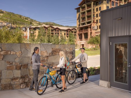Three people with bicycles talking near a building, with mountains and trees in the background on a sunny day.