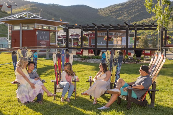 People relaxing in colorful ski-chair seats on grass, with a mountain background and a building nearby.