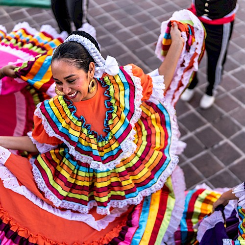 A person dancing in a vibrant, traditional Mexican dress with colorful layers and lace trim, surrounded by others in lively attire.