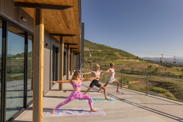 Three people are practicing yoga on a spacious balcony with scenic views of hills under a clear sky.