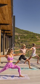Three people are practicing yoga on a balcony with a scenic mountain view in the background under a clear sky.