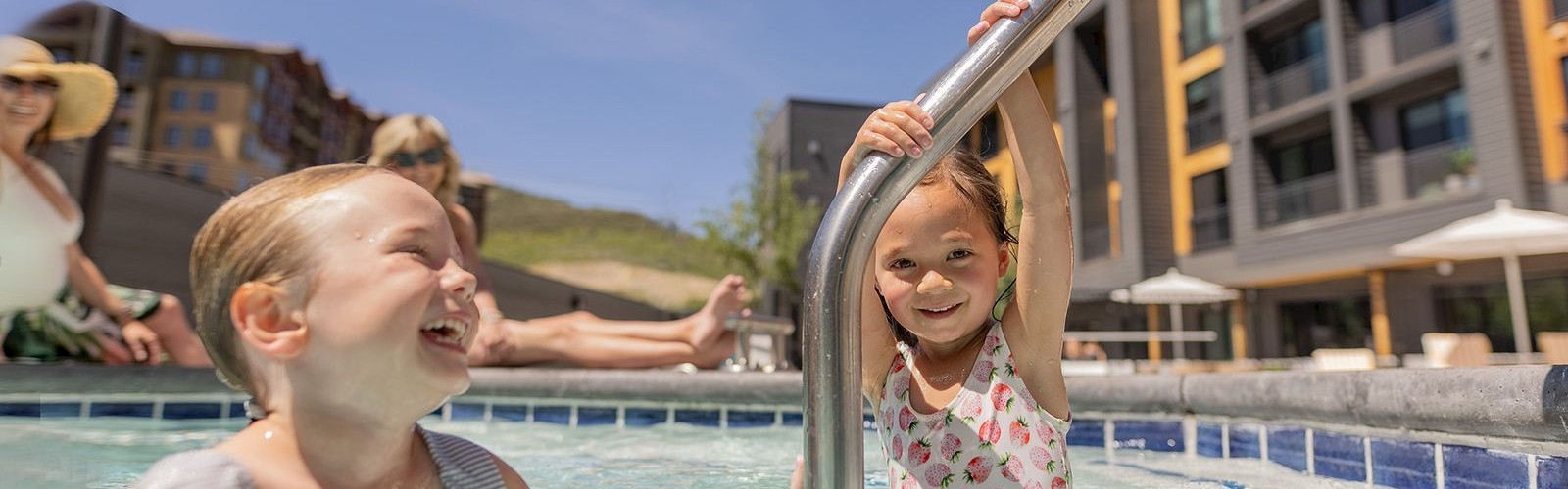 Two kids are happily playing in a pool, with adults relaxing in the background and buildings nearby under a clear sky.