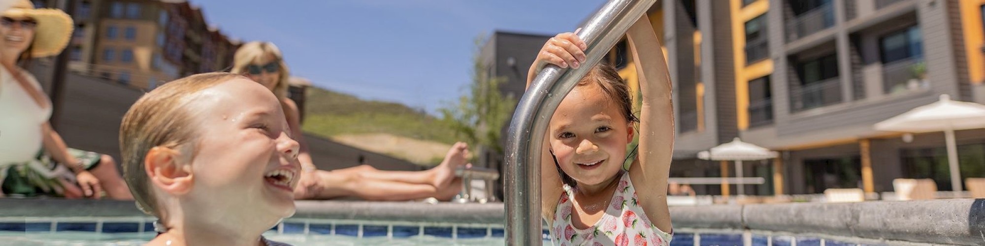 Two kids are happily playing in a pool, with adults relaxing in the background and buildings nearby under a clear sky.