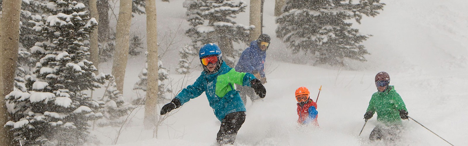 A group of people skiing down a snowy slope surrounded by trees, wearing colorful ski gear and helmets in the winter landscape.
