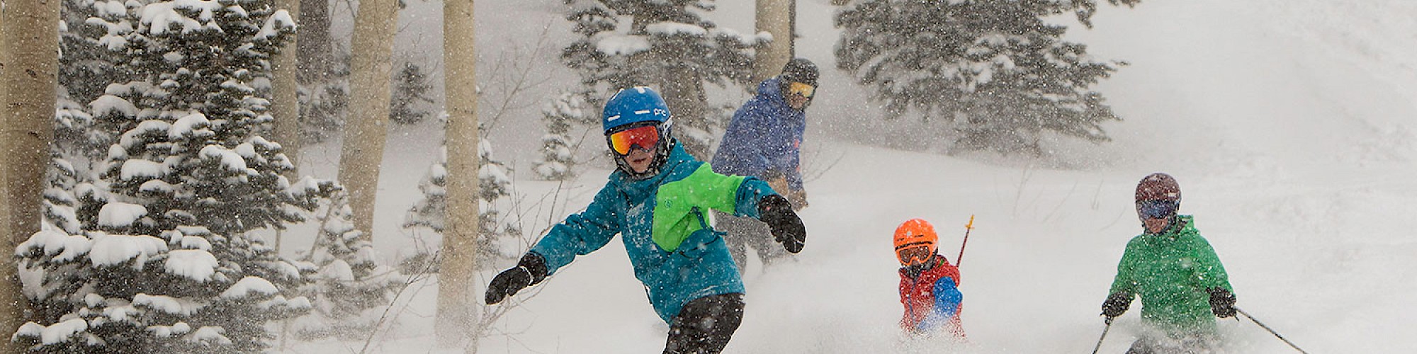 A group of people skiing down a snowy slope surrounded by trees, wearing colorful ski gear and helmets in the winter landscape.