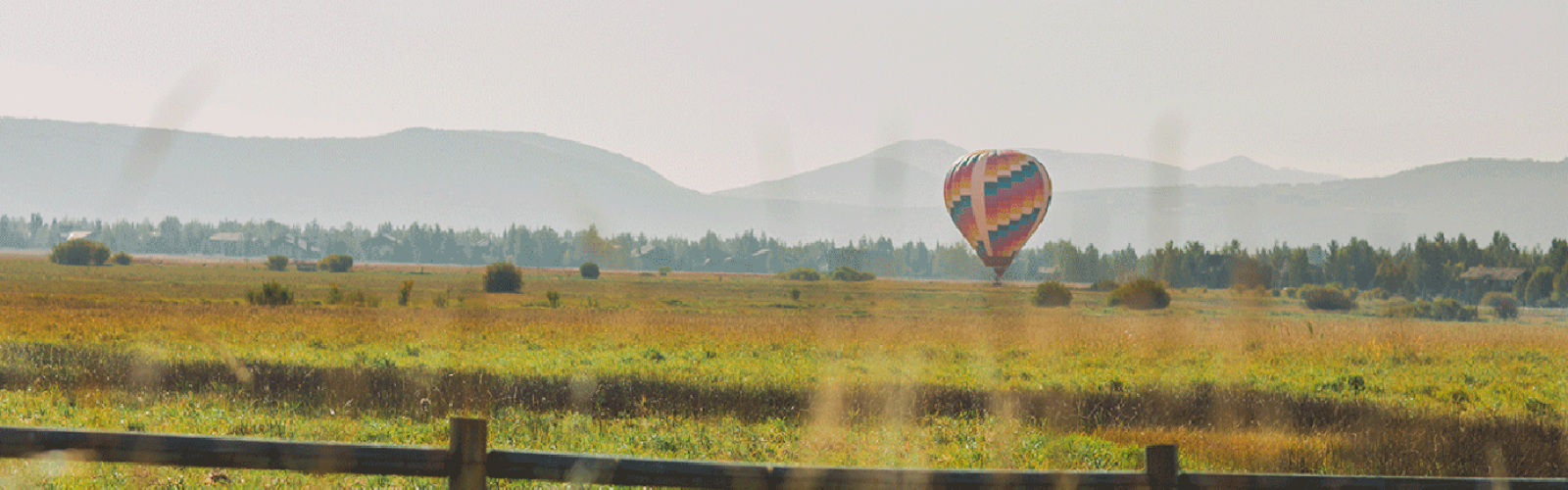 A colorful hot air balloon floats over a scenic meadow, with mountains in the background and a wooden fence in the foreground.