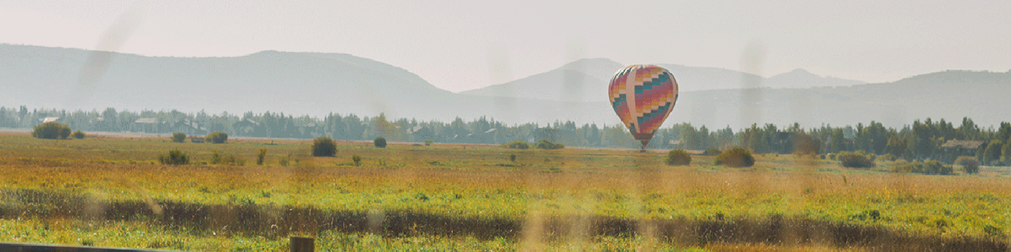 A colorful hot air balloon floats over a scenic meadow, with mountains in the background and a wooden fence in the foreground.