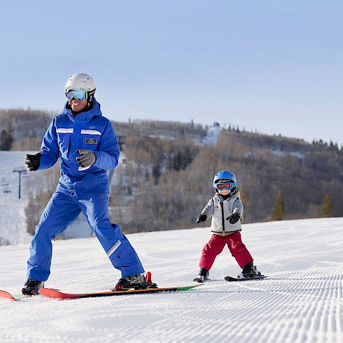 An adult is teaching a child to ski on a snowy slope, both wearing helmets and goggles, with trees and ski lifts in the background.