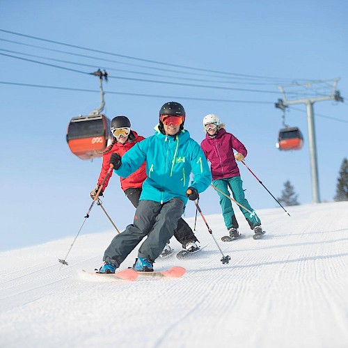 Three people are skiing downhill on a snowy slope with a ski lift in the background, under a blue sky.