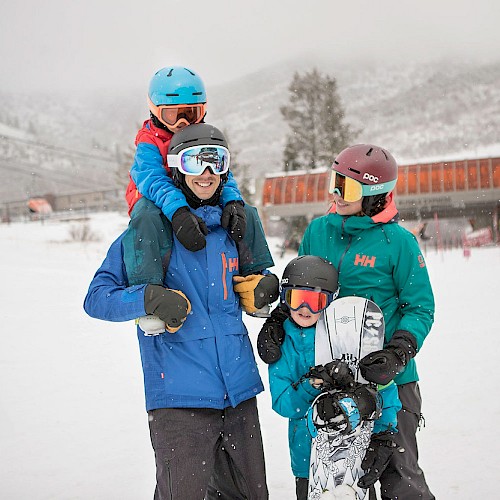 A family of four, dressed in winter gear, poses on a snowy ski slope. The child is on a shoulder. All wear helmets and goggles.